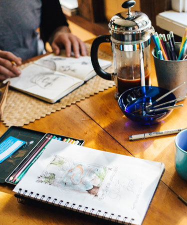 Kreativität ausleben. Foto von einem Holztisch mit Maluntensilien, Sketchblock und Kaffee