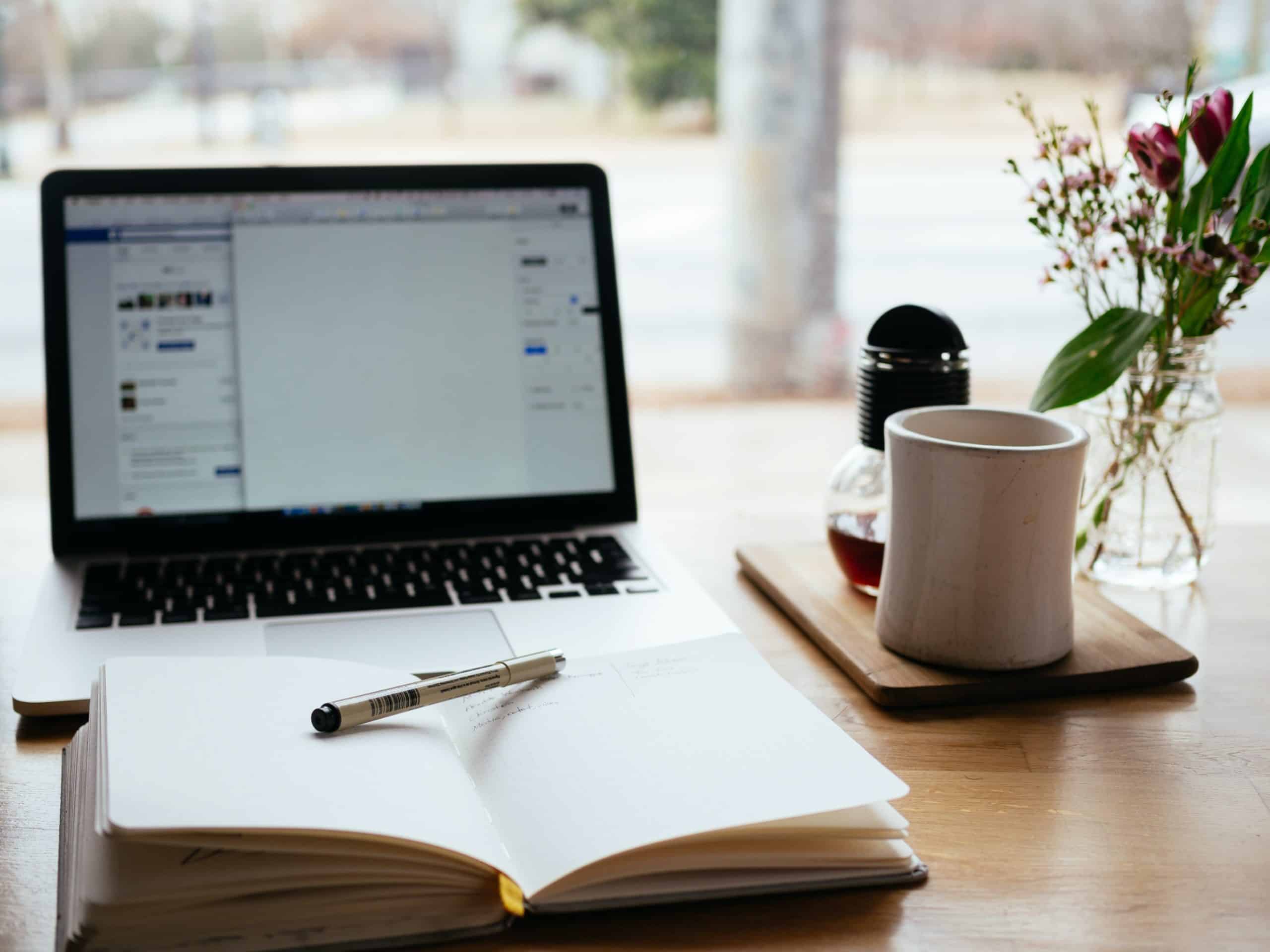 Schreibtisch mit Laptop, Teetasse und -Kanne sowie Blume vor Panoramafenster. Vor dem Laptop liegt ein Notizbuch. Titelbild für die Textarbeit wie etwa das Lektorat.