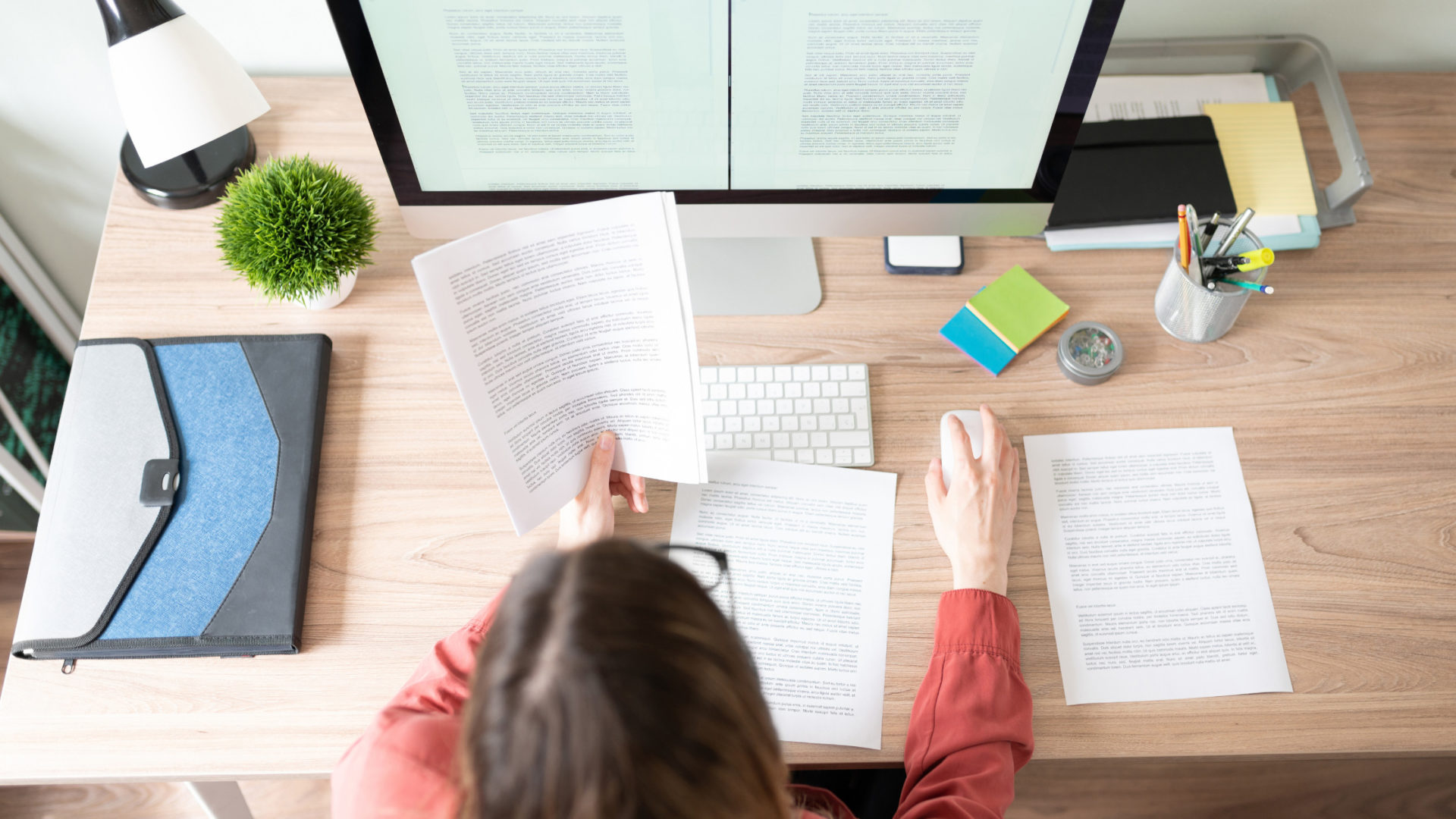 Foto von einer Person, die an einem Schreibtisch sitzt, auf dem einige bedruckte Papiere liegen. Das Foto ist im Rücken der Person, etwas oberhalb des Kopfes aufgenommen worden, sodass man auch den Monitor gut sehen kann. Titelbild für den Blogartikel "Manuskript überarbeiten".